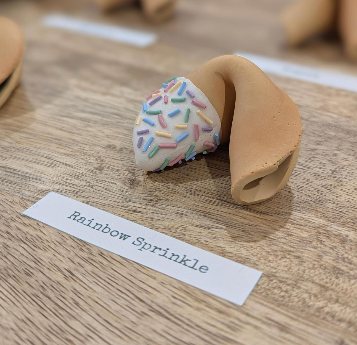 A Rainbow Sprinkle Smart Fortune Cookie, dipped in white with pastel sprinkles, beside its name card